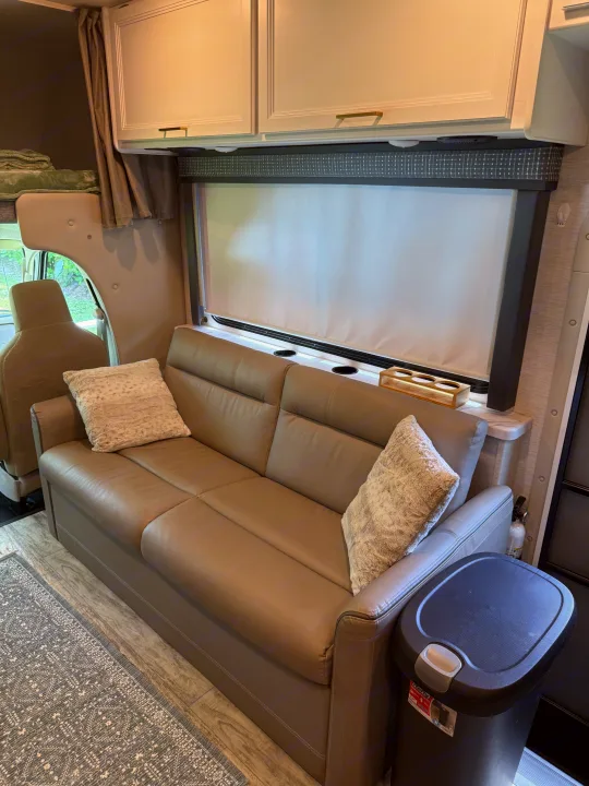 This RV rental's living area features a comfortable taupe sofa, overhead cabinets, and a bright window with a pull-down blind.. Thor Chateau 2025