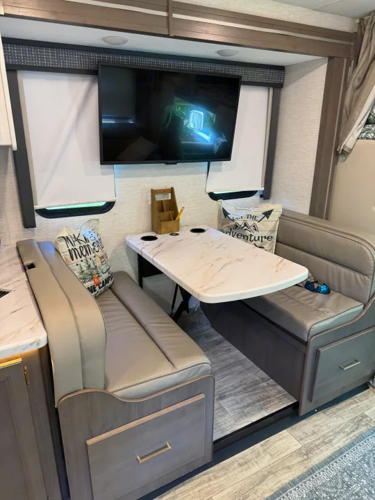 This inviting RV rental dinette features comfy benches, a marble-look table with cup holders, and a wall-mounted TV.. Thor Chateau 2025