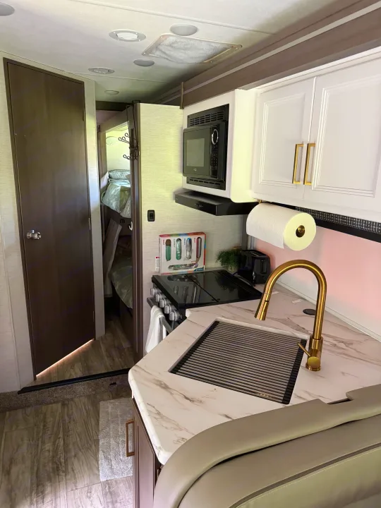 Modern rv rental kitchen: gold faucet, marble counter, white cabinets, and bunk beds visible through a doorway.. Thor Chateau 2025
