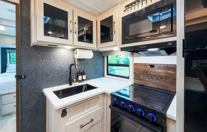 This modern rv rental kitchen features light wood cabinets, black appliances (sink, stove with blue lights, microwave), and a window view.. Forest River Surveyor Legend 2026