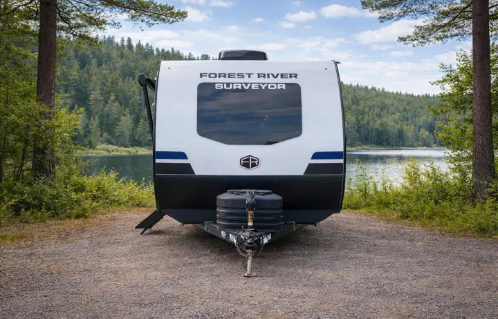 RV rental: A Forest River Surveyor travel trailer is parked on gravel, facing a serene lake and forest. Perfect for outdoor camping.. Forest River Surveyor Legend 2026