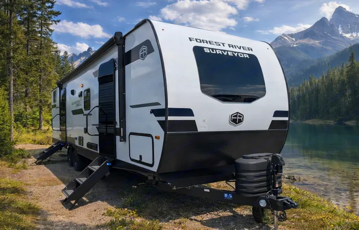 Scenic rv rental: A white Forest River Surveyor travel trailer by a clear lake, surrounded by mountains and pine trees.. Forest River Surveyor Legend 2026