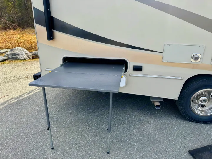 Side of an RV rental showing an extended gray fold-out table, parked on asphalt. A tire, exhaust, and natural background are visible.. Coachmen Leprechaun 260DS Ford 450 2019