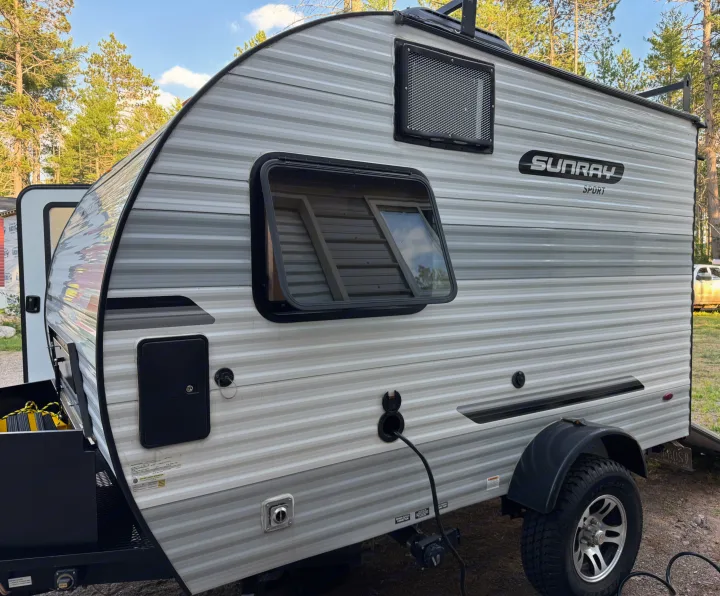 Side view of a grey and white Sunray Sport travel trailer, ideal for an RV rental. Parked outdoors amidst pine trees.. Sunset Park RV SunRay 2022