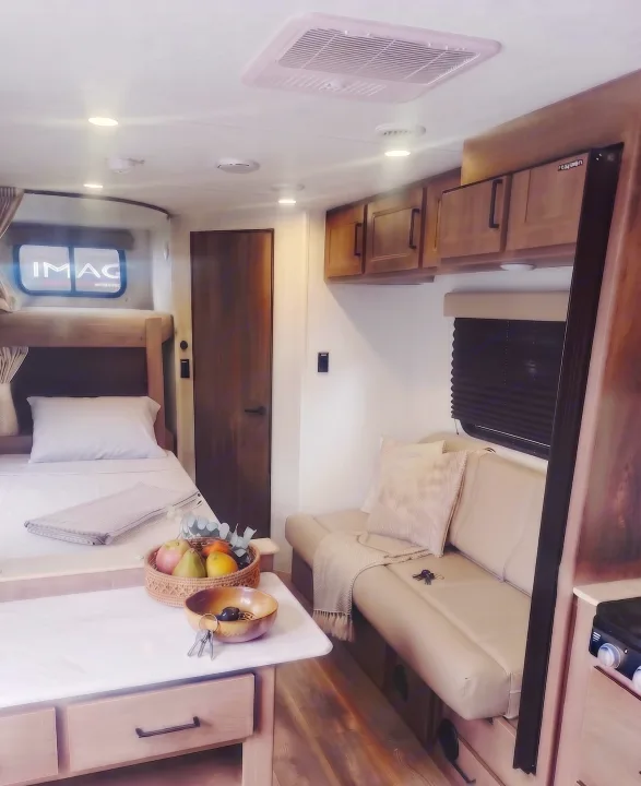 Cozy RV rental interior: a bunk bed with white linen, a light-colored sofa, and a table with fruit and keys.. Grand Design Transcend Xplor 2025