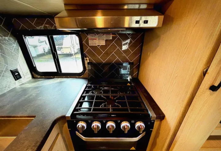 RV rental kitchen: gas stove, oven, dark counter, herringbone backsplash, and window. Compact yet functional for travel.. Coachmen Apex 2022