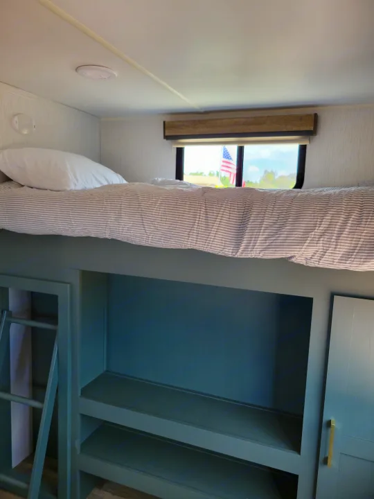 rv rental bunk area with striped bedding, shelves, and a window revealing an American flag.. Heartland RVs Eddie Bauer 2024