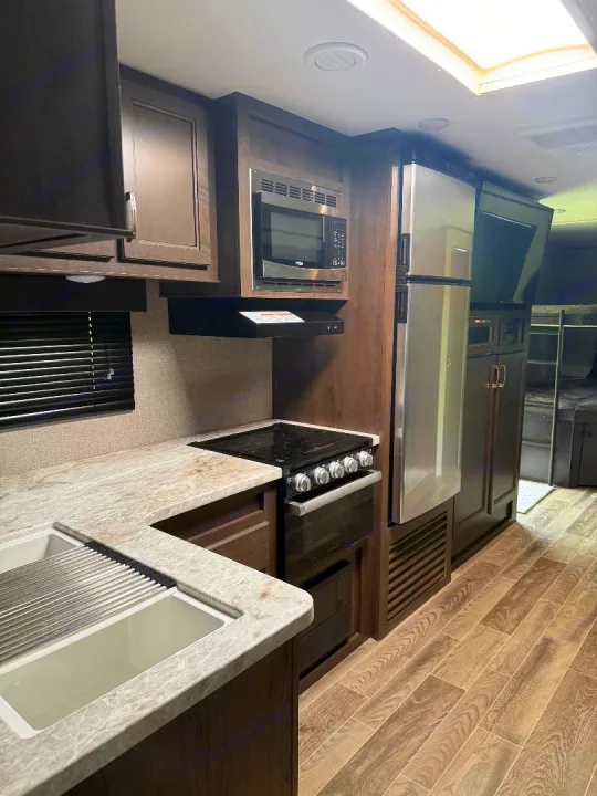 This RV rental kitchen features a modern sink, stovetop, oven, microwave, and refrigerator, all surrounded by stylish wood cabinetry.. Jayco Jay Flight 2021