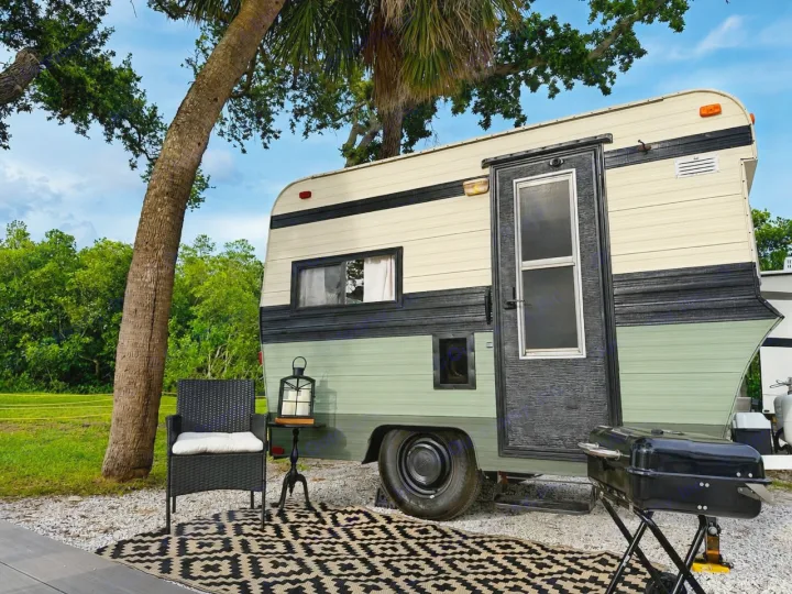 This charming RV rental features a cream and green vintage-style camper, outdoor rug, chair, lantern, and grill setup under a palm tree.. Bell Coaches Bell 1300 1977