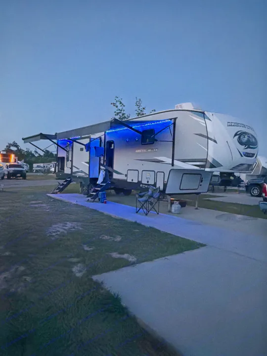 An illuminated RV rental, a fifth-wheel trailer, glows with blue LED lights at a twilight campsite where people relax.. Forest River Artic wolf 2022