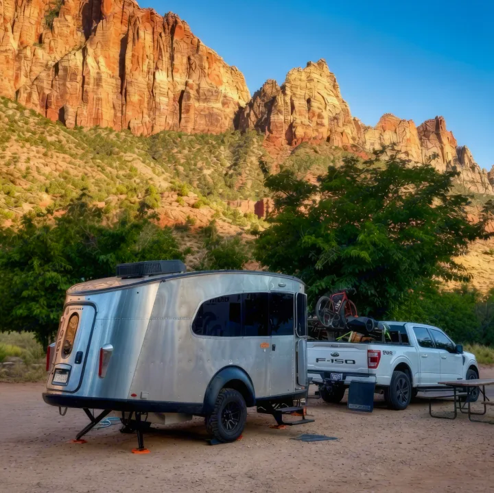 A sleek silver RV rental and white truck are parked at a picturesque campsite with majestic red mountains. Ideal for outdoor adventures.. Airstream Basemamp 2025