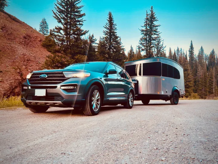 A dark SUV tows a modern silver rv rental camper on a gravel mountain road, ready for adventure among the pines.. Airstream Basemamp 2025