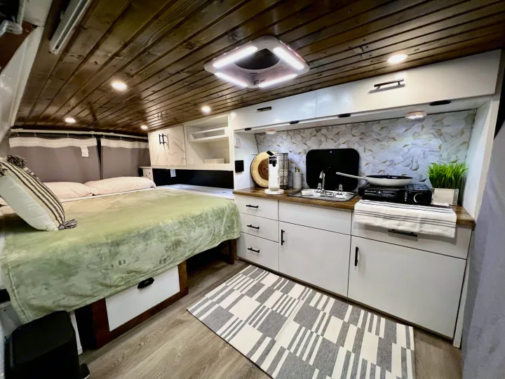 RV rental interior: comfy bed with green blanket, compact kitchen with patterned backsplash, and wood-paneled ceiling.. Ford Ford E350 2005