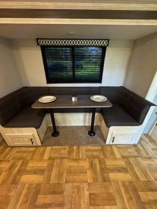 RV rental dinette: U-shaped dark booth seating, gray table with plates, window with blinds, wood-look floor.. Forest River Shasta 2022