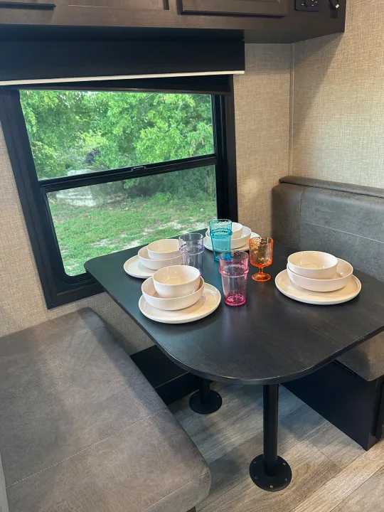 RV rental dining area: dark table set with white bowls/plates, colorful glasses, and a window view of green trees.. Jayco Jay Flight 2023