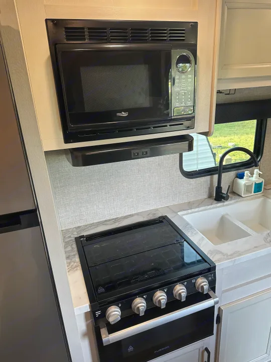 This RV rental kitchen features a black microwave, range hood, Furrion stove with glass cooktop, and a white double sink.. Jayco Jay Flight 2023