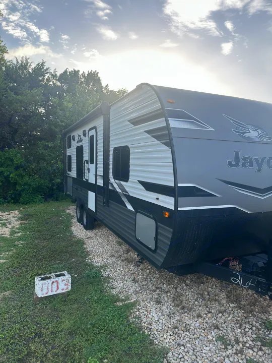 A modern grey and white Jayco rv rental travel trailer is parked on a gravel site surrounded by green trees under a bright sky.. Jayco Jay Flight 2023