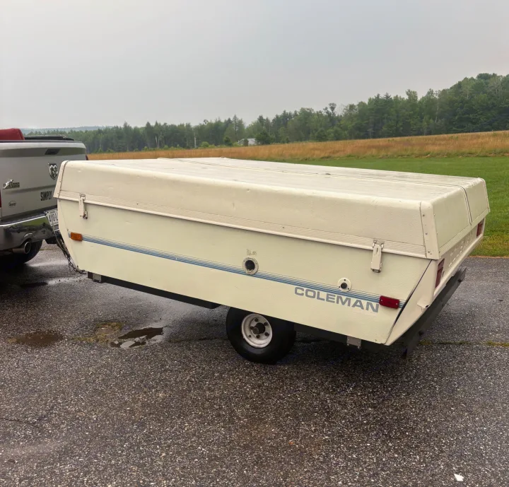 A white Coleman pop-up camper is hitched to a truck on a wet road, perfect for an RV rental adventure through the scenic woods.. Coleman Destiny Roanoke 1991