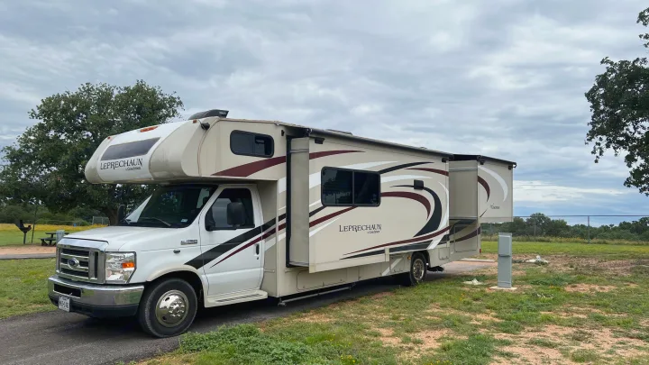 A white and tan Coachmen Leprechaun rv rental, with slide-outs extended, is parked at a grassy campsite under a cloudy sky.. Forest River Coachmen Leprechaun 2020