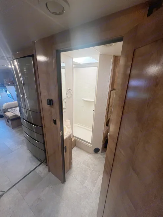 RV rental interior: View into the spacious bathroom with shower and vanity. A modern stainless steel refrigerator and seating are visible.. Forest River Coachmen Leprechaun 2020