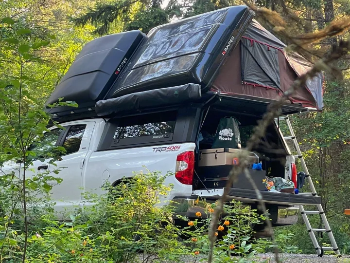 Overland truck with iKamper rooftop tent & ladder, amidst green foliage. An ideal rv rental for outdoor adventures.. Chevrolet Silverado 2024