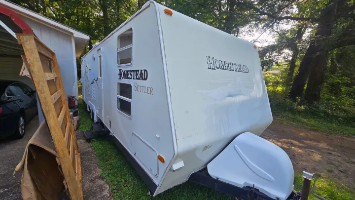 A white Homestead Settler travel trailer, ready for an rv rental. Parked outside, alongside a building and trees.. Starcraft Homestead Settler 2004