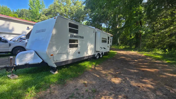 White Homestead Settler RV rental, a travel trailer, parked outdoors next to a truck & dirt path surrounded by lush trees.. Starcraft Homestead Settler 2004