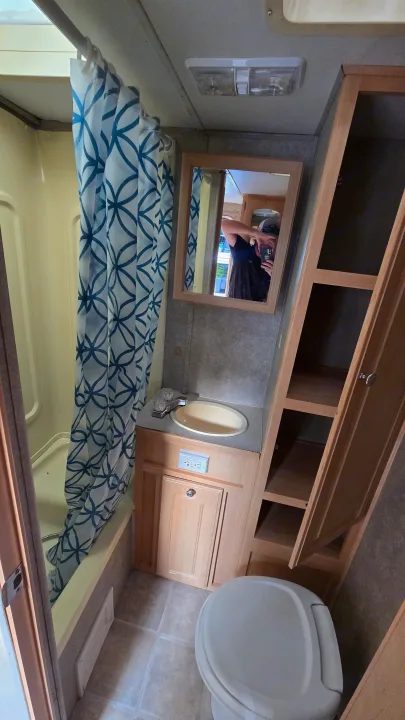 Inside an RV rental: a compact bathroom featuring a shower with a blue pattern curtain, sink, toilet, mirror, and wooden cabinet storage.. Starcraft Homestead Settler 2004