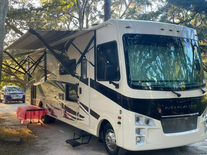 A white & black Coachmen Mirada motorhome rv rental is set up for camping, with its awning extended in a sunny outdoor spot.. Coachmen Mirada 32ss 2020