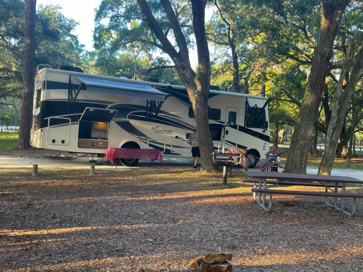 An RV rental, a Coachmen Miranda, is set up at a leafy campsite with its awning out, an outdoor kitchen, and a picnic table nearby.. Coachmen Mirada 32ss 2020