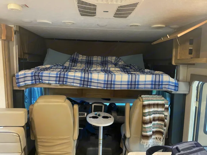 This rv rental features a cozy overhead bed with plaid bedding, visible above the front cockpit and seats.. Coachmen Mirada 32ss 2020