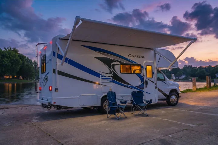 RV rental parked by a tranquil lake at sunset. The 'Chateau' RV has its awning extended and lights on, with chairs set up.. Thor Chateau 2023