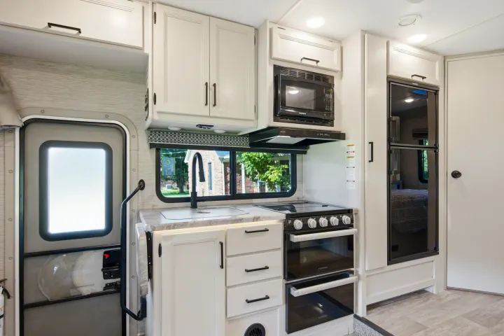 Explore this bright, well-equipped RV rental kitchen featuring white cabinets, modern black appliances, a sink, and an entry door.. Thor Chateau 2023
