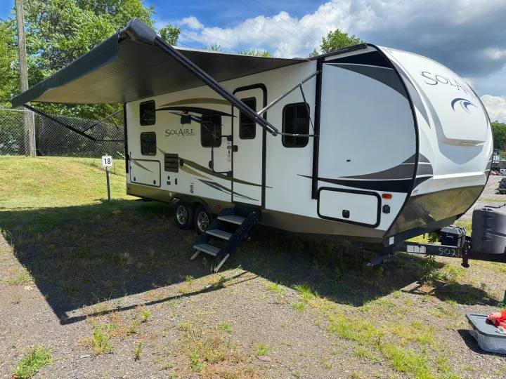An RV rental, a white and black travel trailer with awning extended, parked at sunny campsite 18 on a gravel lot.. Palomino Solaire 240BHS 2018