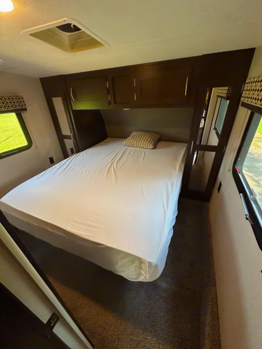 Inside a cozy RV rental bedroom. A neatly made bed is flanked by windows, with overhead storage visible above.. Sportsmen Sportsmen Fifth Wheel 2018