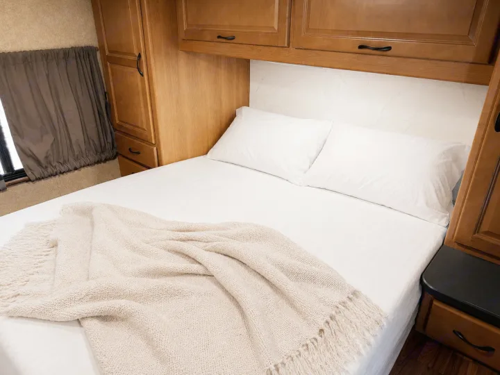 Cozy RV rental bedroom features a comfy bed with white linens, a knitted blanket, and ample wood storage for your travels.. Thor Majestic 2017