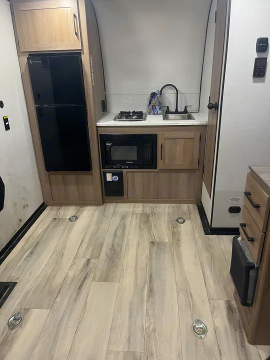 Modern rv rental kitchen interior: black fridge, stove, sink, microwave, light wood cabinets, and flooring with tie-downs.. Keystone Coleman 13T 2026