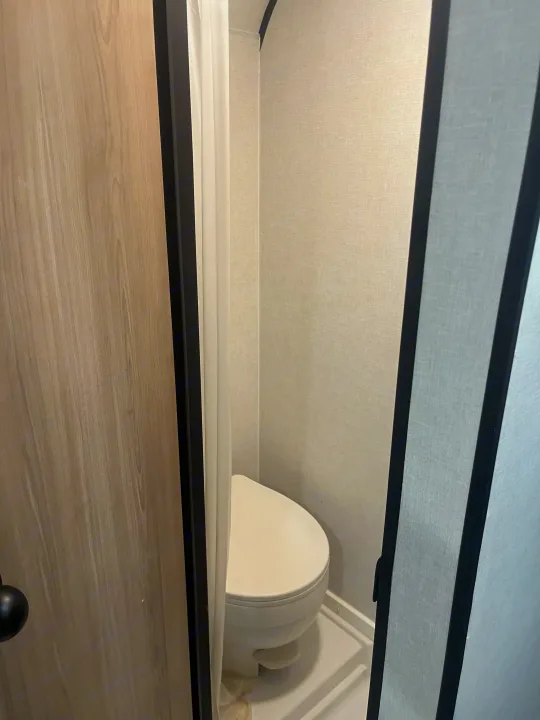 Inside an RV rental, a compact bathroom features a white toilet, a white shower curtain, and light textured walls.. Keystone Coleman 13T 2026
