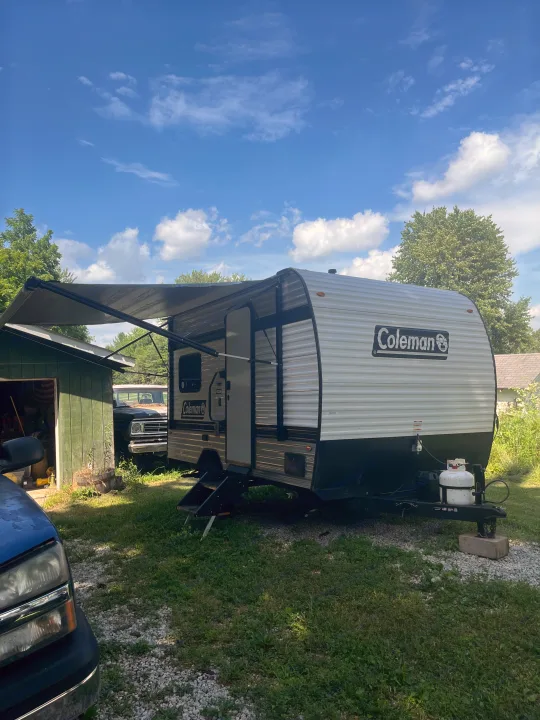 A modern white and black Coleman travel trailer with its awning extended, parked on gravel and grass near a green shed. An excellent rv rental option.. Keystone Coleman 13T 2026