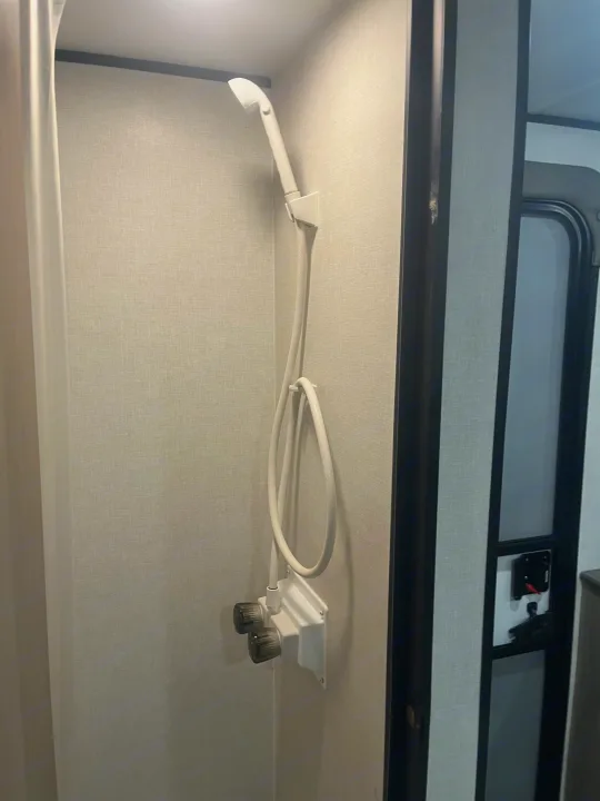 RV rental shower: Compact white handheld shower with dual knobs in a textured grey stall, ideal for travel.. Keystone Coleman 13T 2026