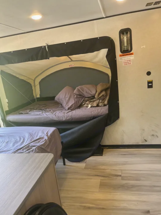 RV rental interior: A pop-up camper bed with bedding and an EXIT sign. A warning label is on the wall, and another bed sits below.. Keystone Coleman 13T 2026
