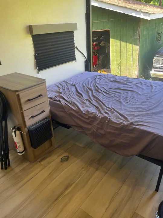 RV rental bedroom: Bed, nightstand with fire extinguisher, pleated blind, and outdoor view of a shed and ATV.. Keystone Coleman 13T 2026