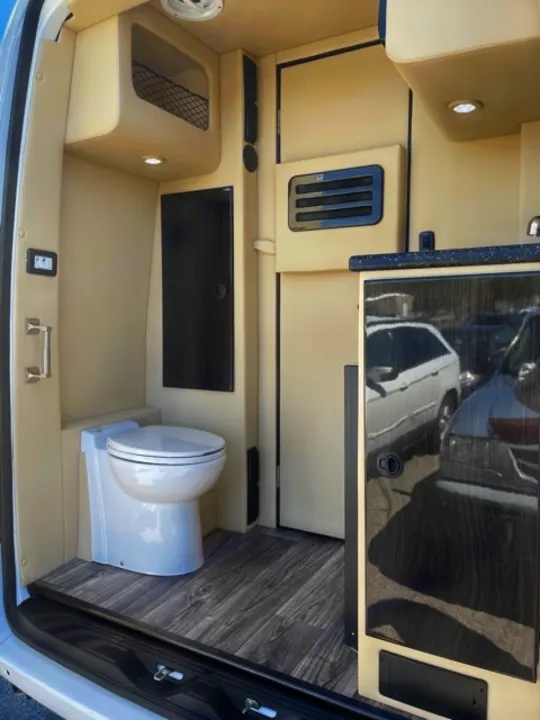 Inside this RV rental: A compact, modern toilet on wood-look flooring, with beige walls, black cabinets, and overhead storage.. Mercedes-Benz 4x4 Sprinter mwb 2020