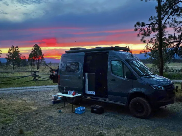 RV rental camper van parked at sunset for a camping adventure. The side door is open, with a table set up outside.. Winnebago Revel 2021