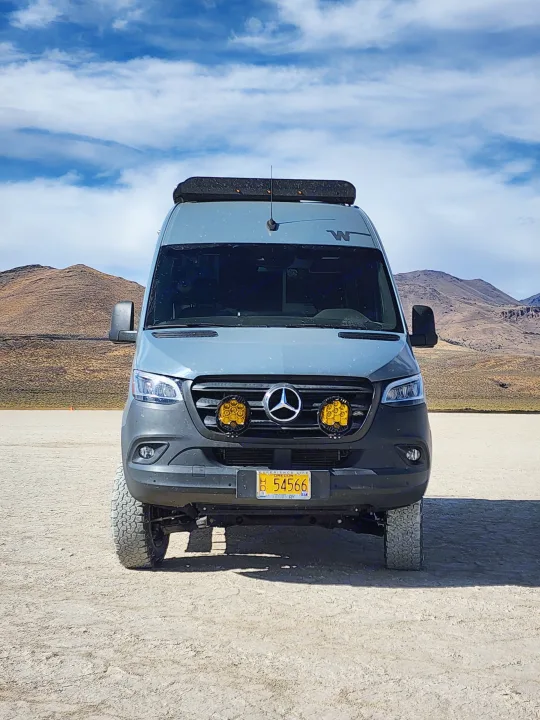 Adventurous grey Mercedes Sprinter rv rental van with off-road lights & roof rack on a desert playa under cloudy skies.. Winnebago Revel 2021