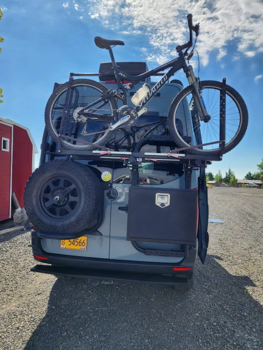 Rear view of an rv rental van with a Specialized mountain bike on a bike rack, spare tire, and storage box, ready for adventure.. Winnebago Revel 2021