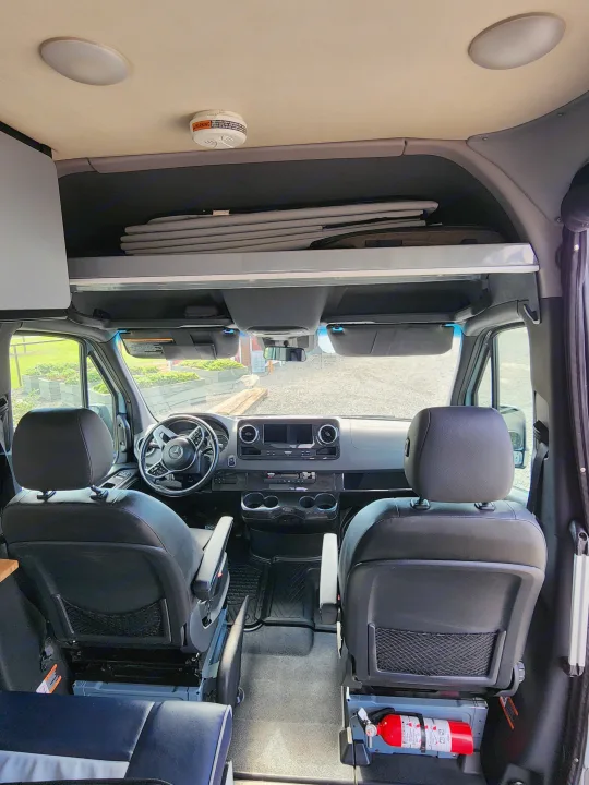 RV rental interior: front cockpit with Mercedes dashboard, spacious overhead storage, and crucial fire extinguisher by the seat.. Winnebago Revel 2021