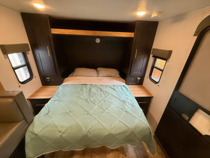Cozy RV rental bedroom featuring a green quilted bed, dark cabinets, wooden nightstands, and windows.. Keystone RV Springdale 2023