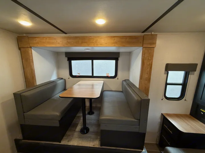 Bright RV rental dinette with grey benches, a wooden table, and windows. Ideal dining space in this well-appointed camper interior.. Keystone RV Springdale 2023