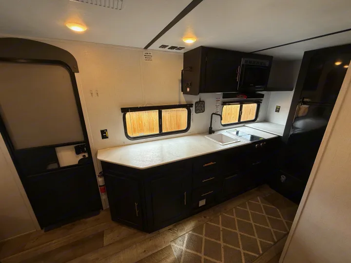 Inside an RV rental, this compact kitchen features black cabinets, white countertops, a sink, stovetop, microwave, and refrigerator.. Keystone RV Springdale 2023